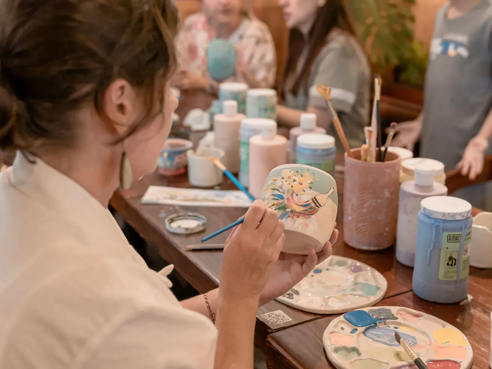 Friends and family painting ceramic cups around a table covered in paints and brushes.