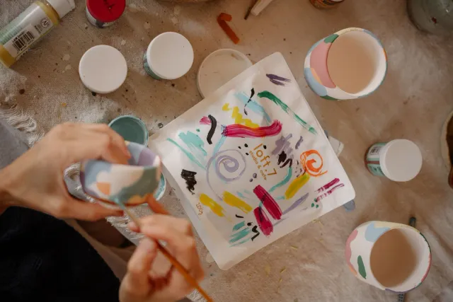 Hands painting a small ceramic cup beside a palette of bright colors, seen from above.