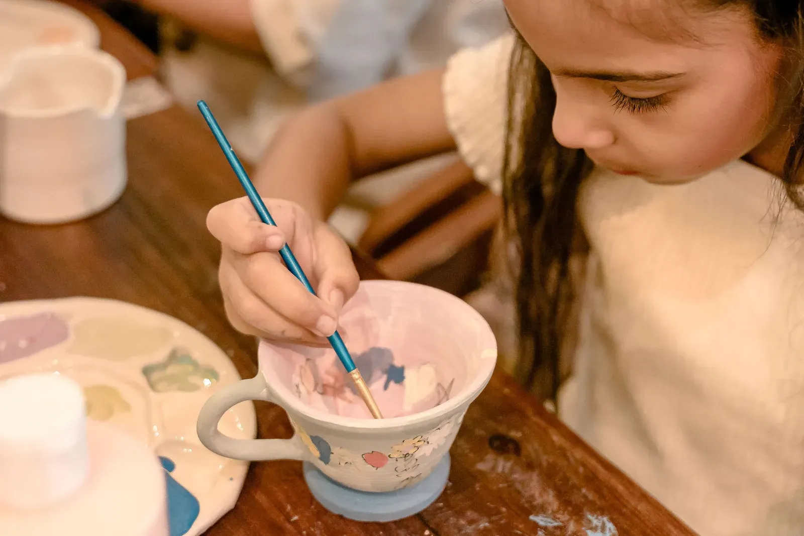A girl painting the inside of a ceramic cup at a workshop table.