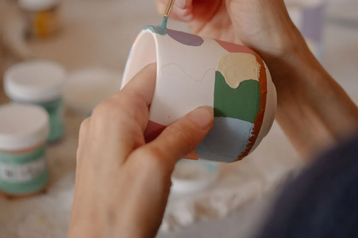 Hands painting colorful shapes on a ceramic cup.